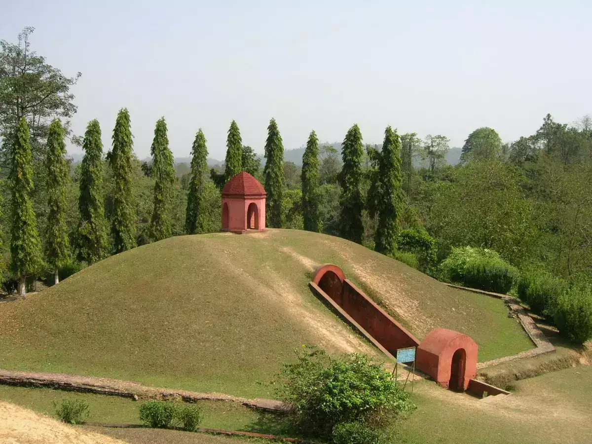 Image of a historic site featuring a red mausoleum atop a mound with a long, arched entrance. The mound is surrounded by lush greenery and tall, evenly-spaced trees. A sign is visible near the entrance. The sky is clear, and the foliage appears verdant.