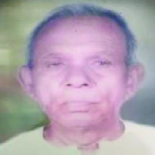 A close-up photograph of an elderly person with short hair and a neutral expression, wearing what appears to be a light-colored shirt. The background is blurred with green and yellow tones.